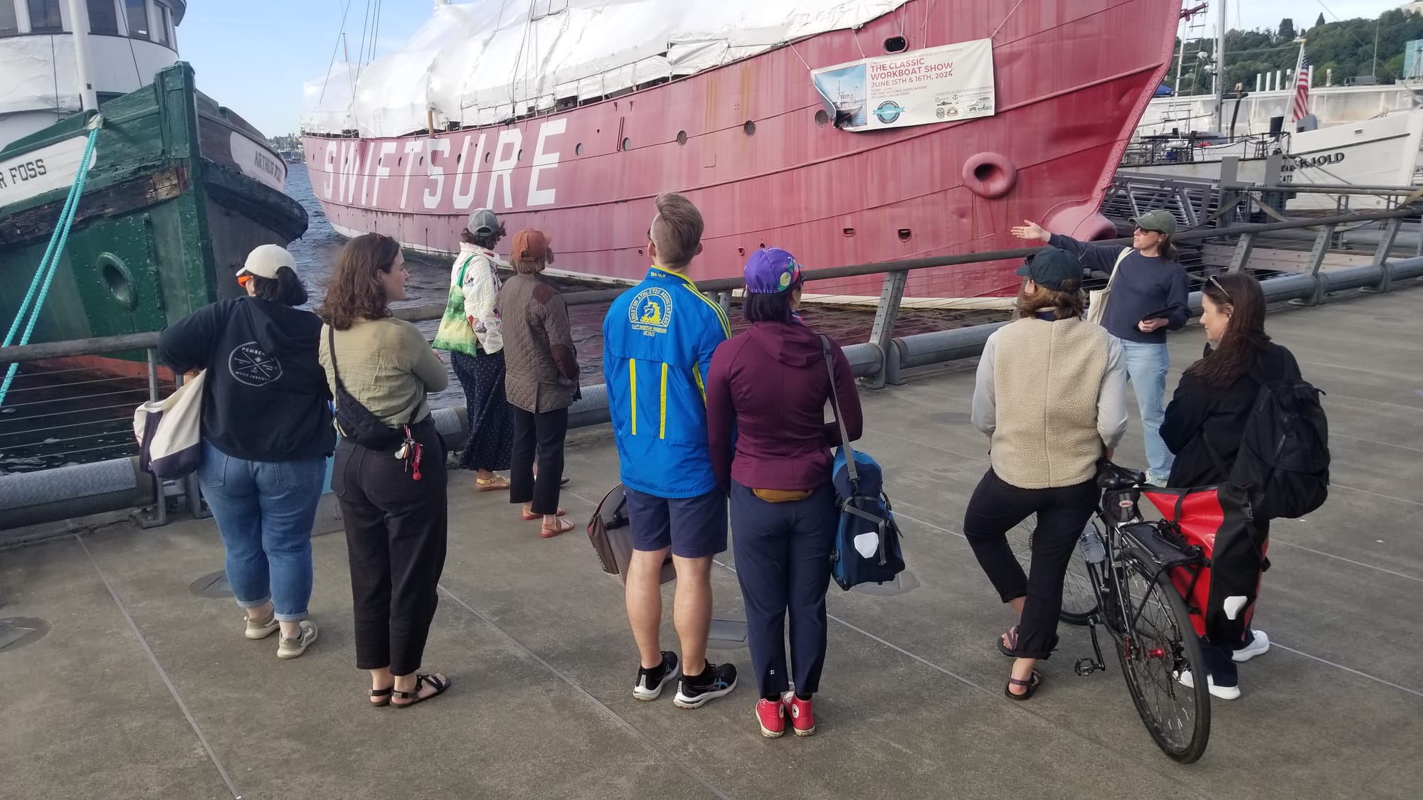 A group of people standing on a dock next to ships listen to a woman talk.