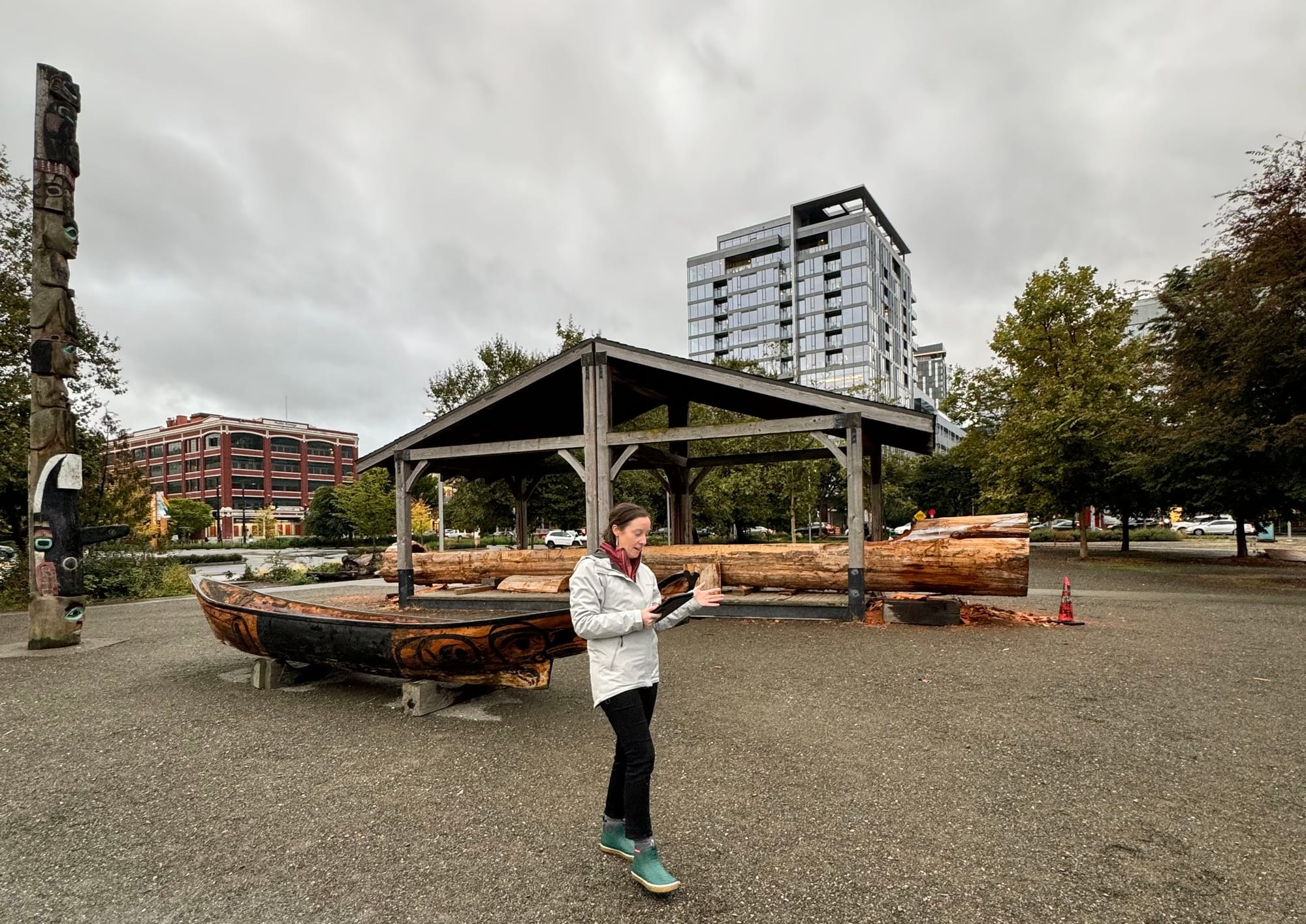 A woman in a rainjacket and boots reads from an iPad standing in front of a wood carved canoe.