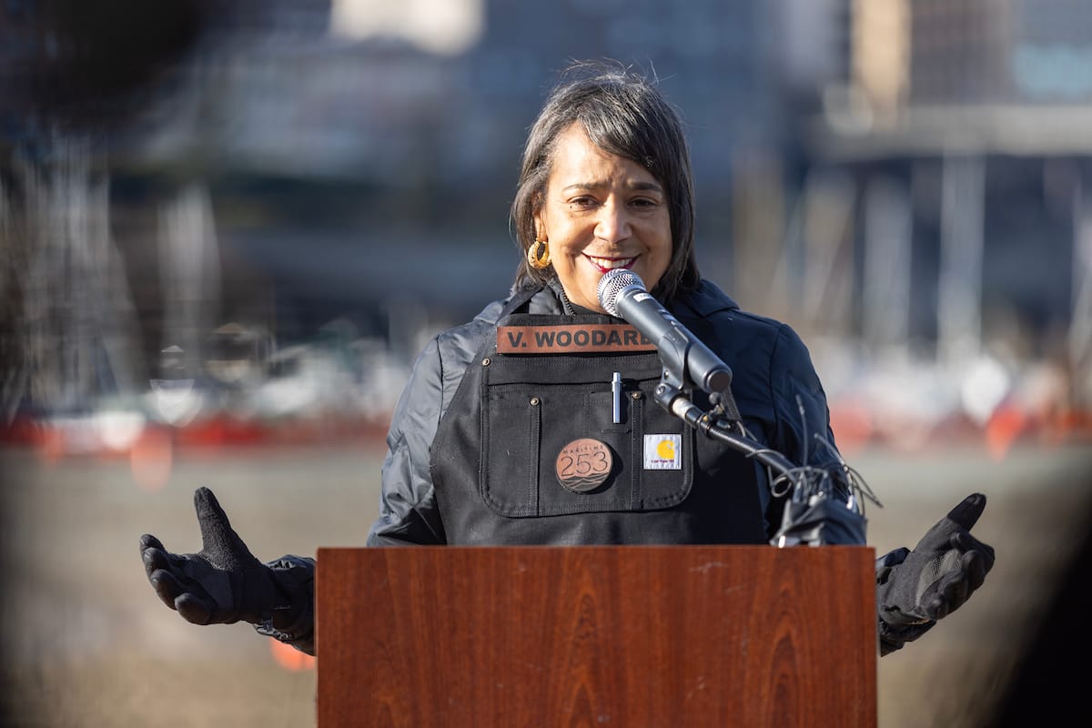 A woman wearing a black work apron speaks at a podium.