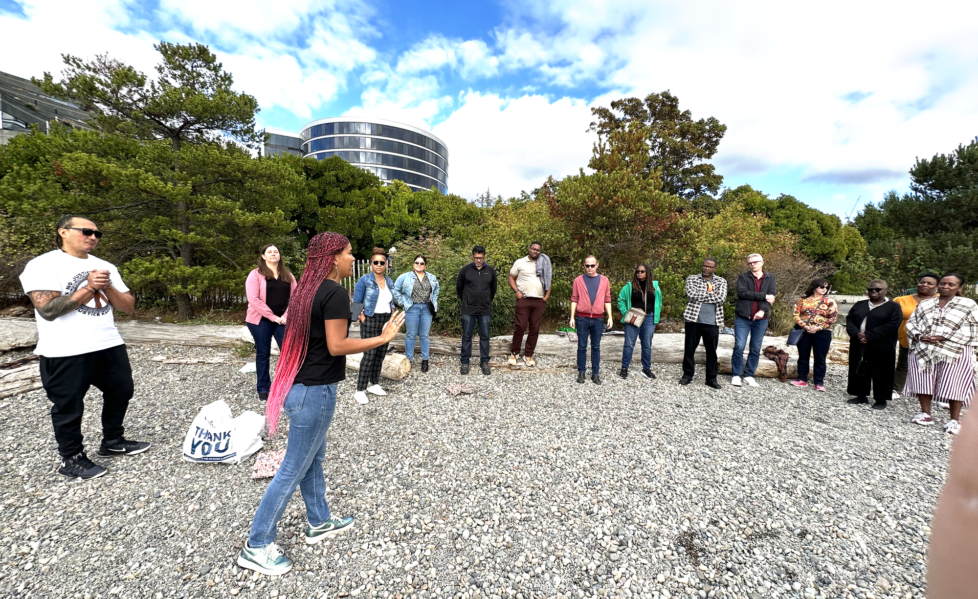 A group of people stand in a half circle on a rocky beach listening to a woman with long pink hair.