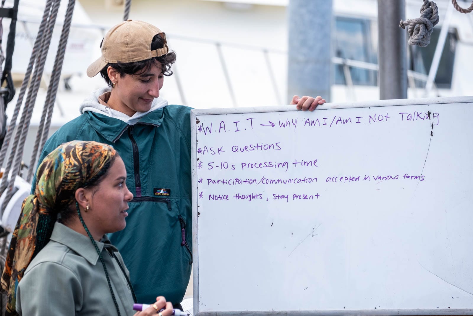 Two people look at a whiteboard with instructions, boats in the background.