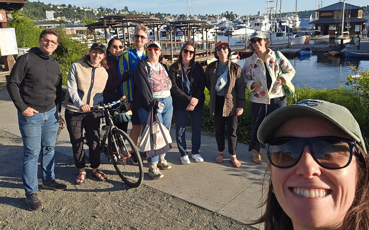 A woman takes a selfie with a group of people behind her on a sunny day with water and boats in the background.