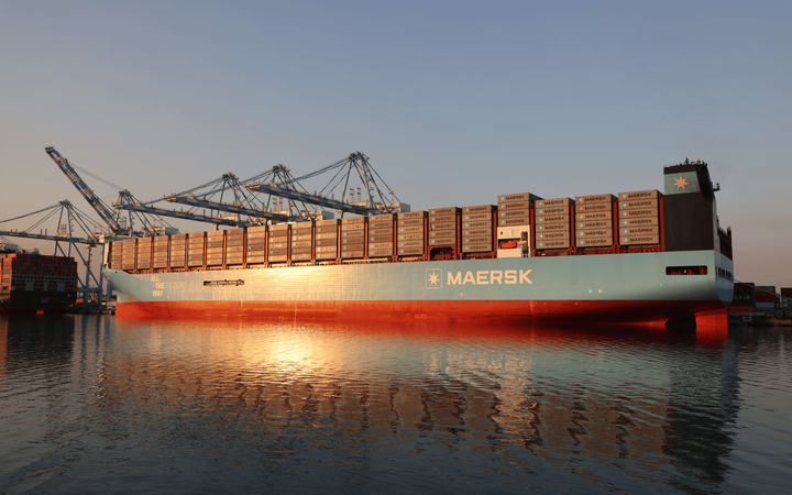 A large blue ship filled with containers at a dock with cranes in the background.