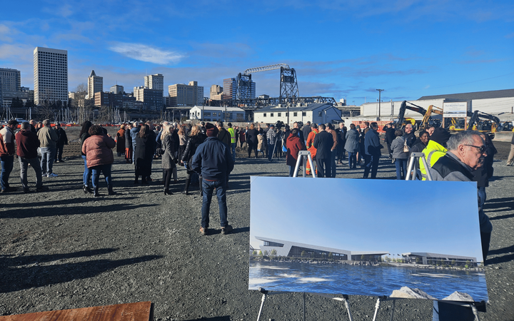 A crowd of people in a gravel lot with an architect's rendering of a new modern building in the foreground.