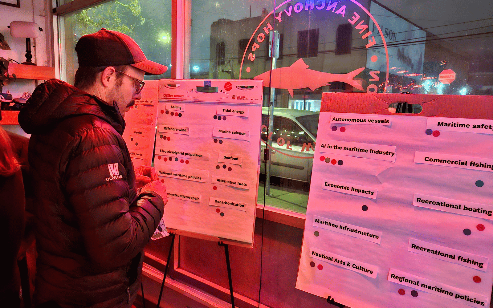 A man in a black puffy jacket and baseball cap looks at posters with text and round stickers.